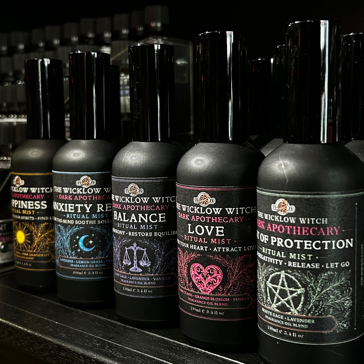 Six dark bottles of Wicklow Witch apothecary ritual mists arranged on a dark shelf, each with a different illustrated label including sun, moon, scales, heart, and pentagram designs.