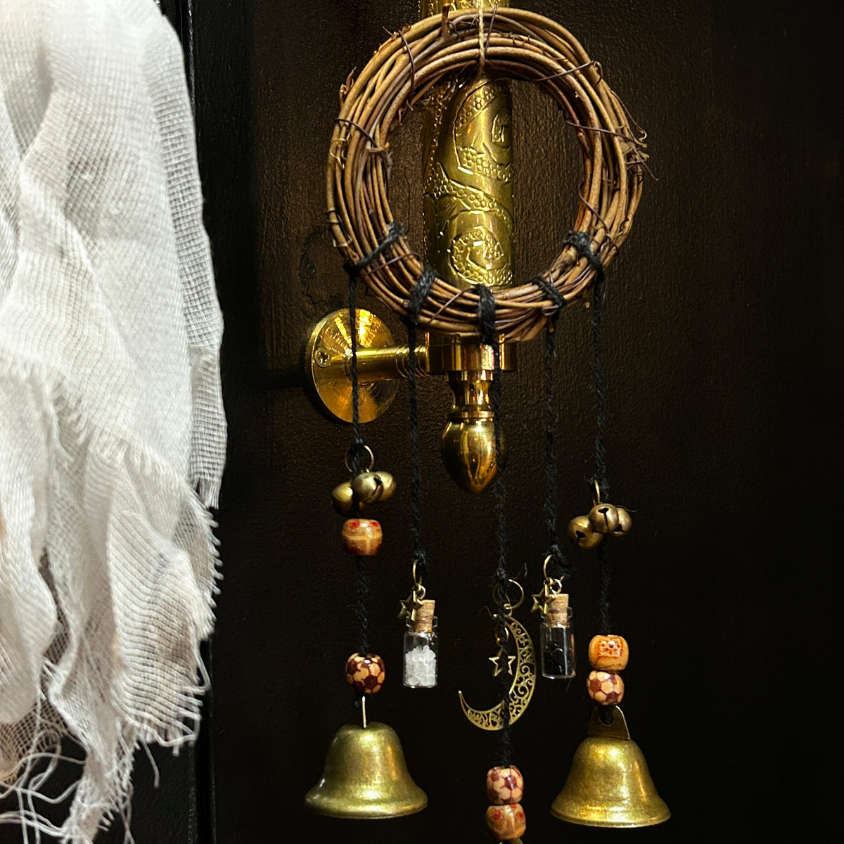A decorative hanging ornament set with three small brass bells, wooden beads, and a woven circular wreath, suspended from a brass door handle on a dark door. A white gauze ghost decoration hangs at the left edge.