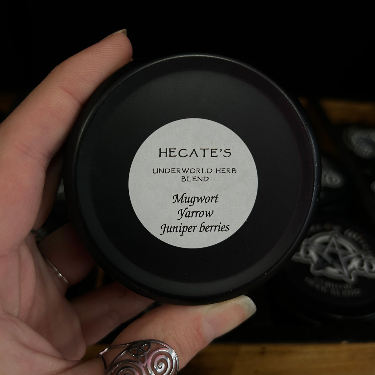 A hand holding a round black tin of Hecate's Underworld Herb Blend Mugwort Yarrow Juniper berries, with a white circular label on the lid.