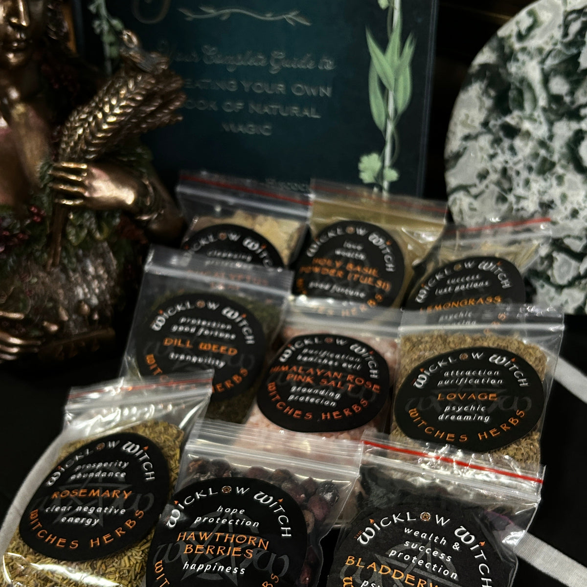 Several small clear plastic bags filled with dried herbs are arranged on a dark tabletop. Each bag has a round black label with orange text reading 'Wicklow Witch' and the name of a herb such as Rosemary, Hawthorn, Bladderwrack, and Cat's Eye.