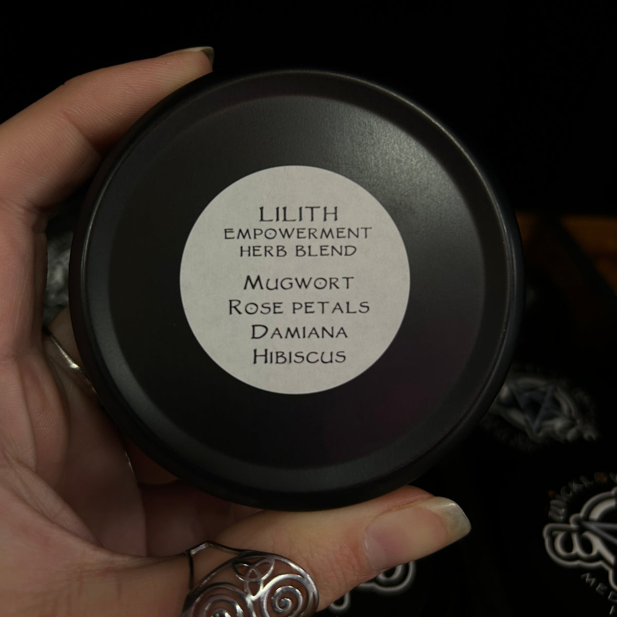 A hand holding a round black tin labeled 'Lilith Empowerment Herb Blend' against a dark background.