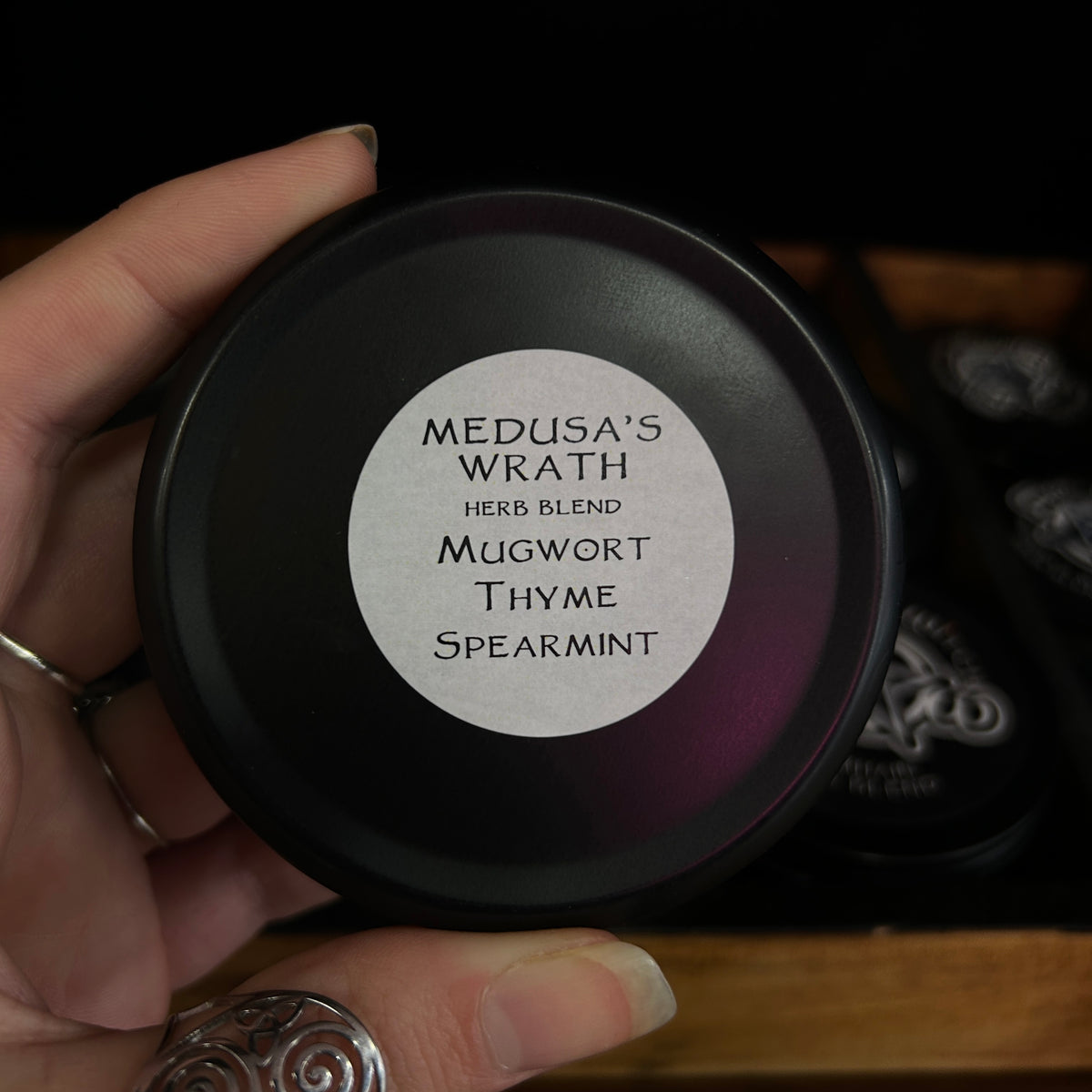 A hand holding a round black tin of 'Medusa's Wrath Herb Blend' mugwort thyme spearmint candle, with a silver label on the lid, set against a dark background with other tins.