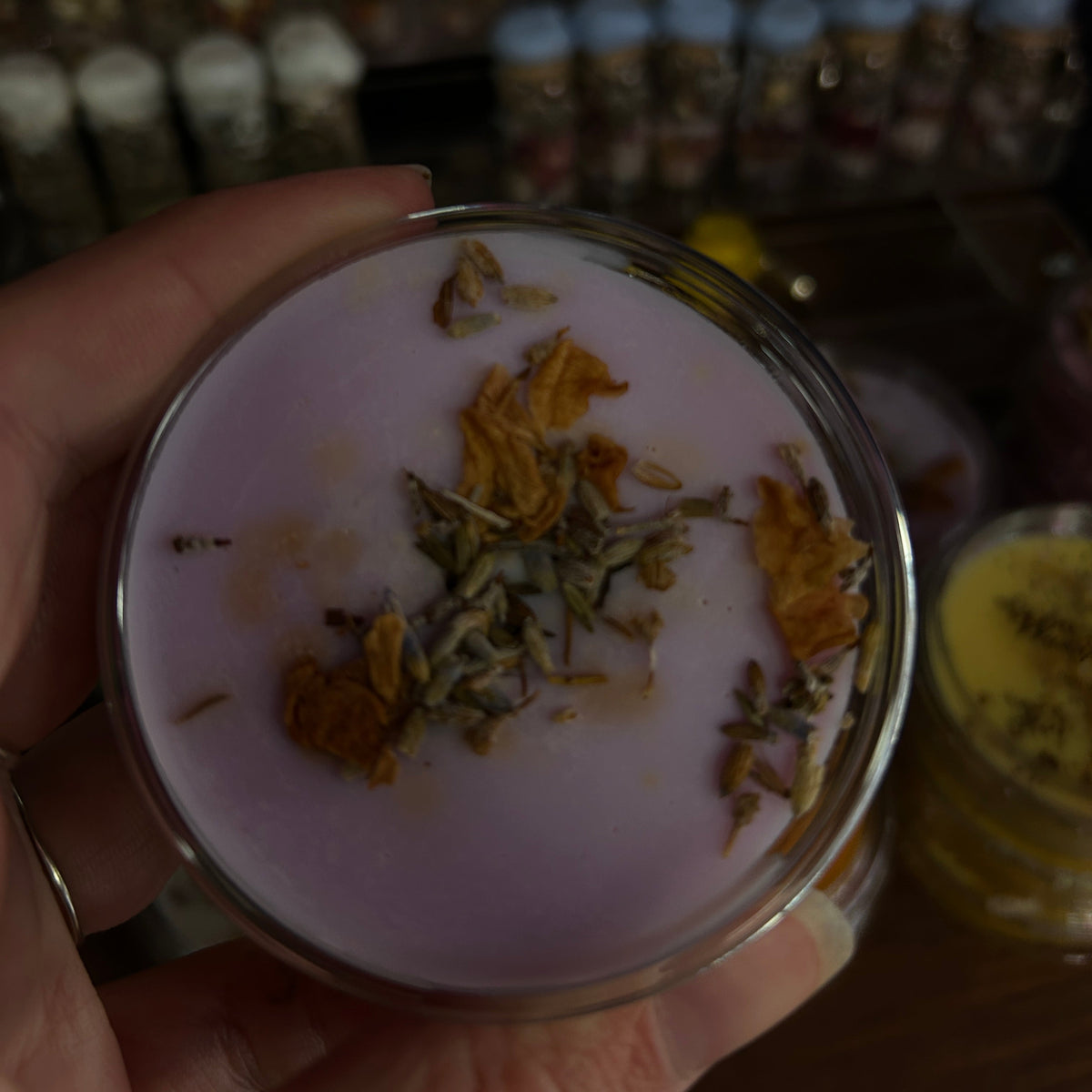A hand holding a small round jar filled with pale lavender-colored wax topped with dried flower petals, with a blurred background of other small jars and bottles.