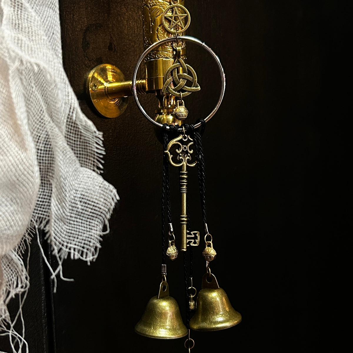 A decorative hanging ornament with three small gold bells, a black cord, pentagram and triquetra charm and ornate key-shaped charms.