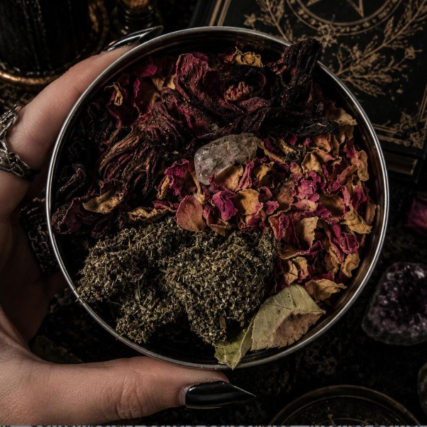 Hand holding a metal tin filled with dried herbs and stones on a dark background