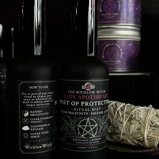 Two black spray bottles from The Wicklow Witch on a dark shelf, with one labeled 'Dark Apothecary Mist of Protection' in the foreground and purple-labeled jars behind it, plus a white dried sage bundle on the right.