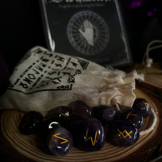 A set of dark purple amethyst oracle stones with gold engraved symbols, displayed on a wooden slice next to a beige drawstring pouch and a framed 'The Book of Divination' card deck box.