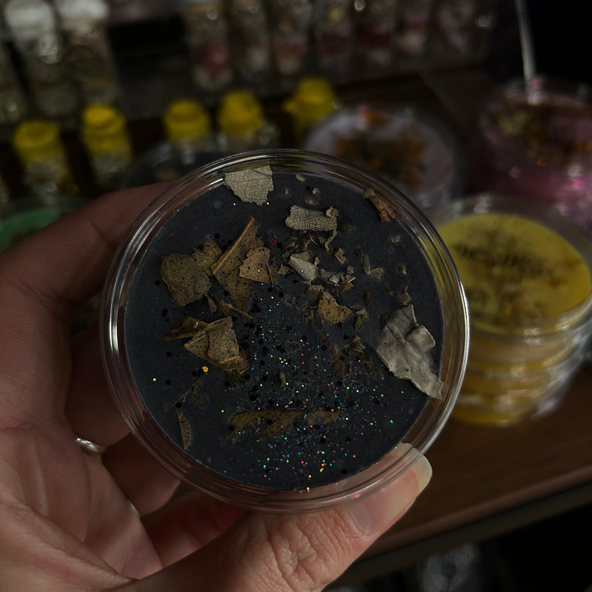 A hand holds a small round clear container filled with a dark charcoal gray cosmetic or wax product, topped with dried botanical flakes and fine glitter. The background is a dimly lit, cluttered shelf with other jars and containers.