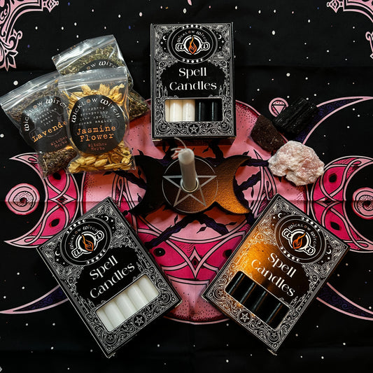 A flat lay of Spell Candles products on a black starry cloth with pink occult symbols. Visible items include three boxed Spell Candles packs, two clear bags of dried botanicals labeled Lavender and Jasmine Flower, a pentacle holder with a white candle, a pink crystal, and a black crystal.