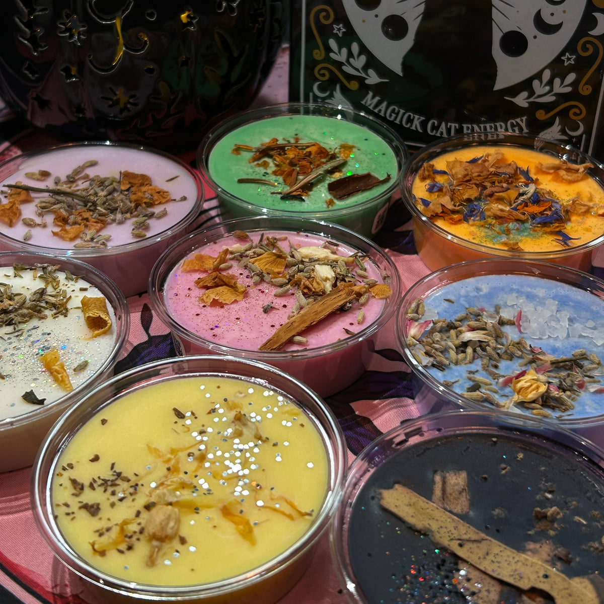 Six colorful wax candles in clear glass jars arranged on a pink tray, with dried botanical toppings. A black cauldron and a 'Book of Spells' card are visible in the background.