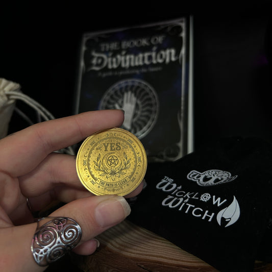 A hand holds a gold-colored divination coin with a 'YES' symbol, with 'The Book of Divination' and a black pouch branded 'The Wicklow Witch' in the background.