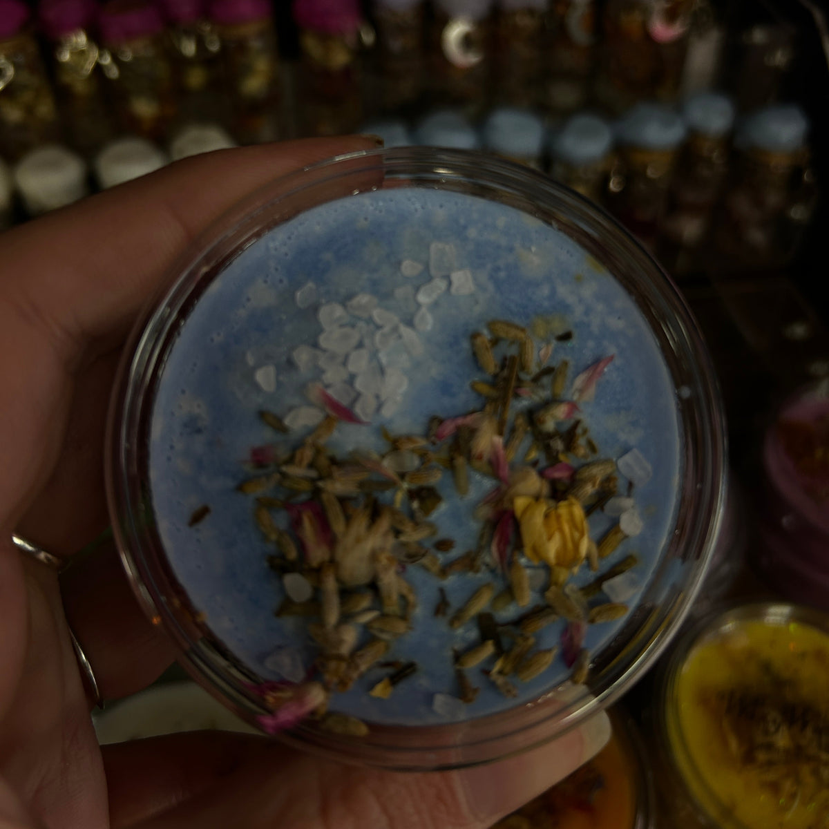 A hand holding a round clear plastic jar filled with a light blue wax melt topped with dried botanicals, and small pink and yellow flower pieces. The background is dark and filled with blurred bottles of similar products.