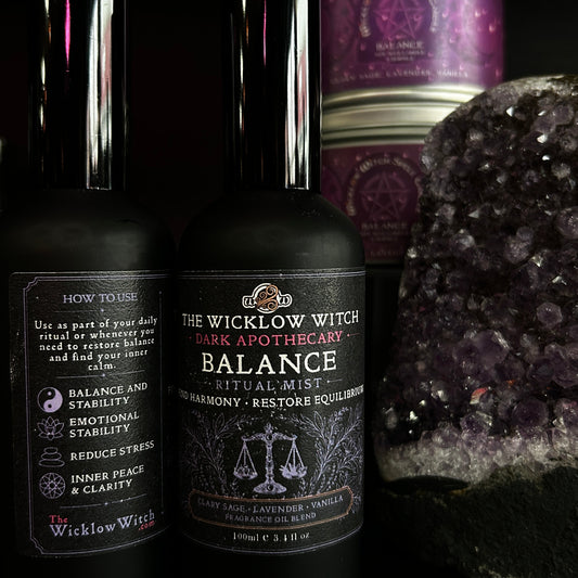 Two black bottles of Wicklow Witch ritual liquids, including 'Balance' and 'Dark Apothecary', displayed alongside a purple amethyst crystal cluster and a purple tin canister on a black background.