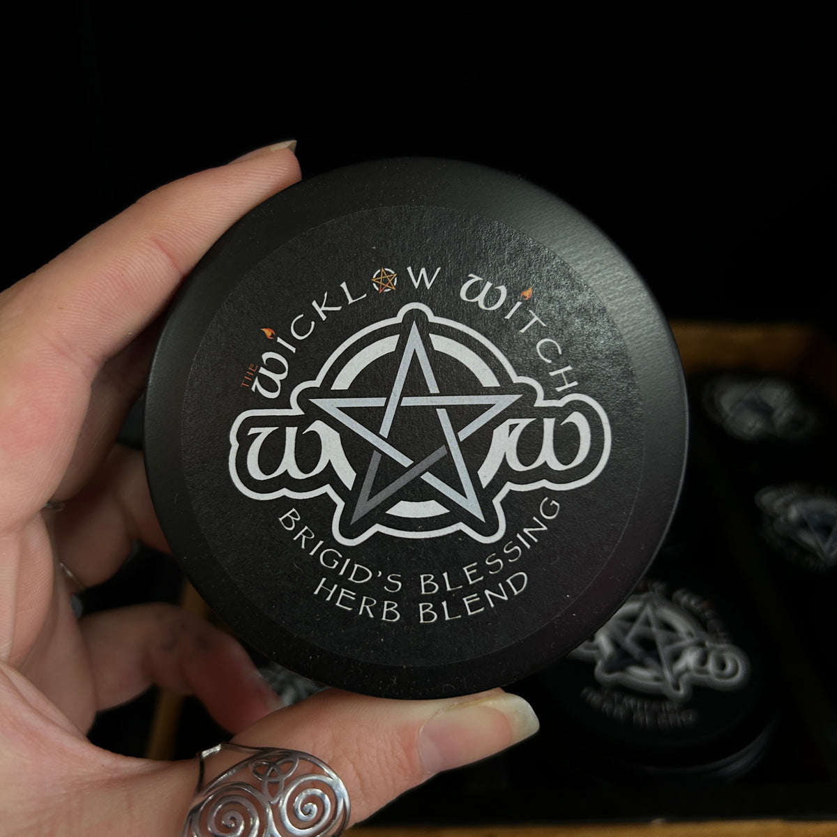 A hand holding a round black tin labeled 'Wicklow Witch Brigid's Blessing Herb Blend' with a white pentagram logo, against a dark background with other matching tins visible.