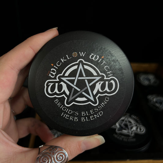 A hand holding a round black tin labeled 'Wicklow Witch Brigid's Blessing Herb Blend' with a white pentagram logo, against a dark background with other matching tins visible.