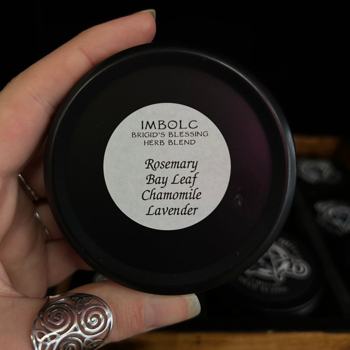 A hand holding a round black tin of Imbolc Bricid's Blessing Herb Blend, with a white circular label listing Rosemary, Bay Leaf, Chamomile, and Lavender. The background is dark with other blurred tins.