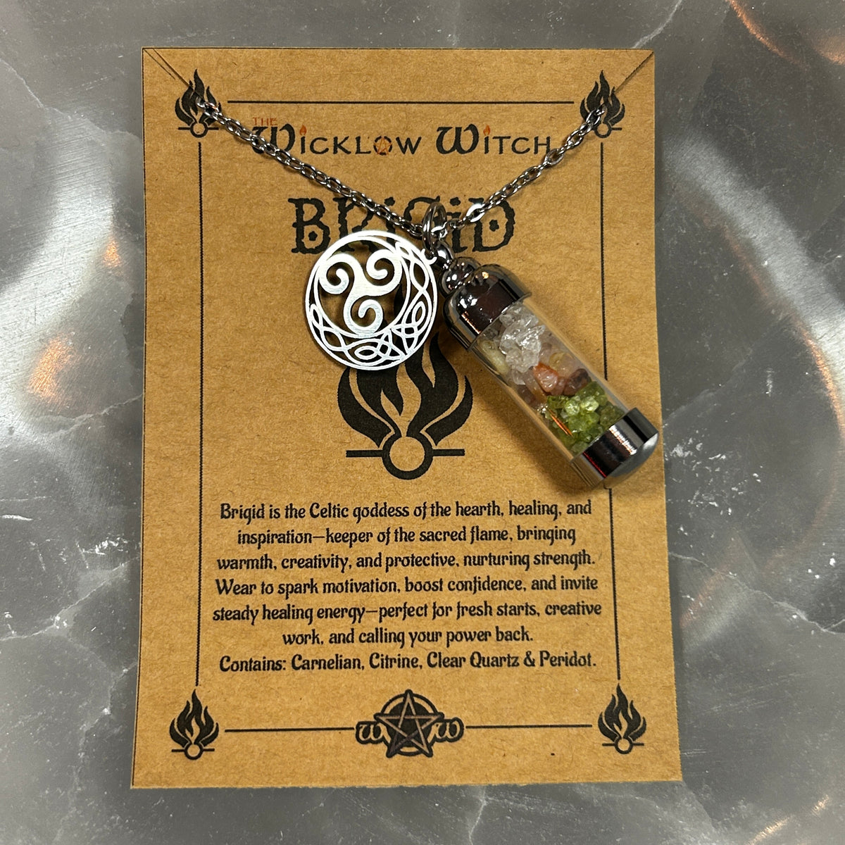 A small glass vial pendant necklace with a silver Celtic knot ball charm, displayed on a brown kraft card labeled 'Brigid' with text about the Wicklow Witch and the contents of the vial, photographed on a metallic surface.