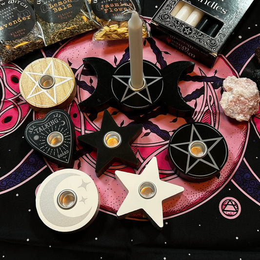 A collection of wooden pentacle candle holders in various shapes (moon, star, heart, circle) in black, white, and beige, arranged on a pink and black mystical cloth alongside raw crystals and packaged incense.