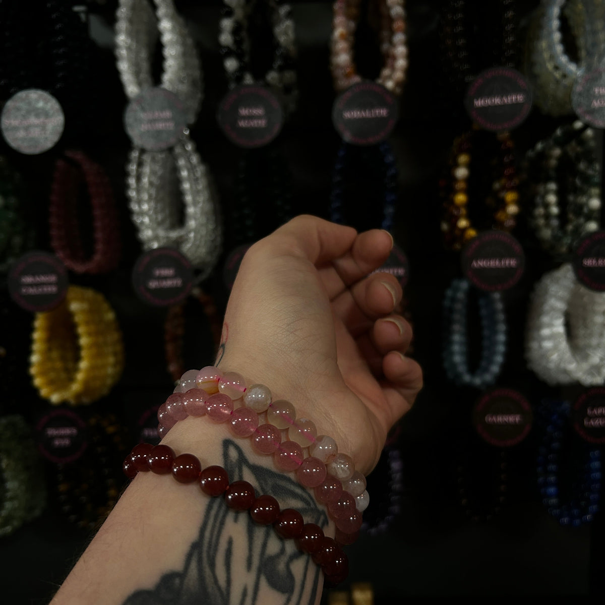 A close-up of a tattooed hand wearing three beaded bracelets, with a display of many colorful crystal bead bracelets hanging in the background.