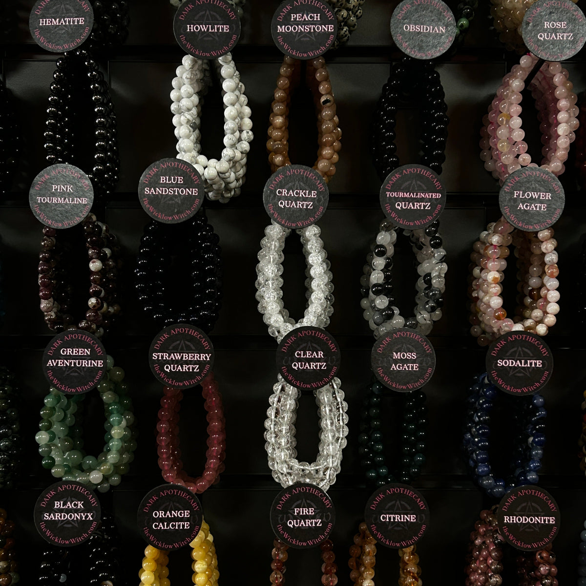 A display of multiple beaded crystal necklaces arranged in rows on a dark background, each labeled with a circular tag showing the stone name such as hematite, howlite, obsidian, turquoise, clear quartz, green aventurine, smoky quartz, hand agate, bodalite, yellow tiger's eye, orange calcite, citrine, and amethyst.