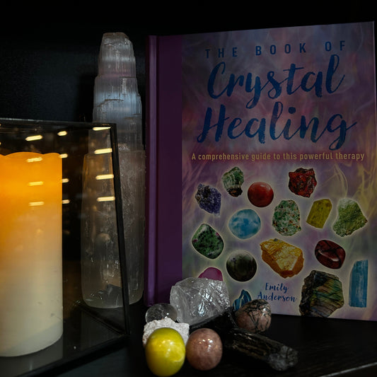 The Book of Crystal Healing by Emily Anderson displayed on a shelf next to a glowing candle, a clear quartz tower, and various crystals and stones.