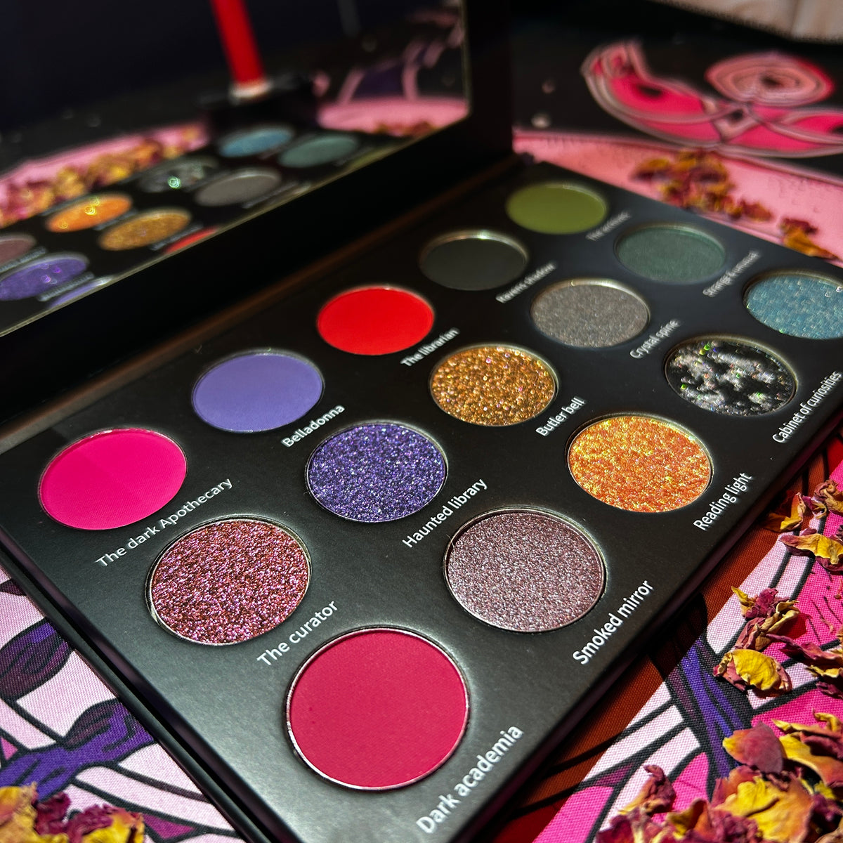 An open makeup palette showing 15 round eyeshadow pans in assorted glitter and matte shades, photographed close-up on a colorful printed surface with dried flowers around it.