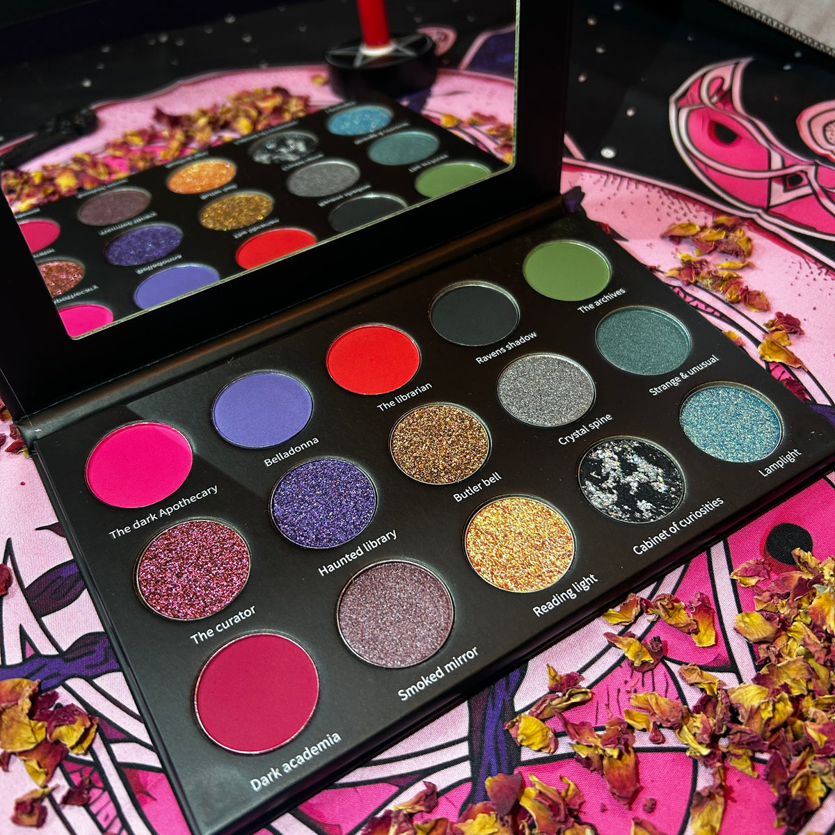 An open 15-pan eyeshadow palette displayed on a pink patterned surface with dried flower petals. The black palette contains a mix of matte, shimmer, and glitter shades in pink, purple, red, green, gold, and blue, with some shades labeled and a mirror visible in the lid.