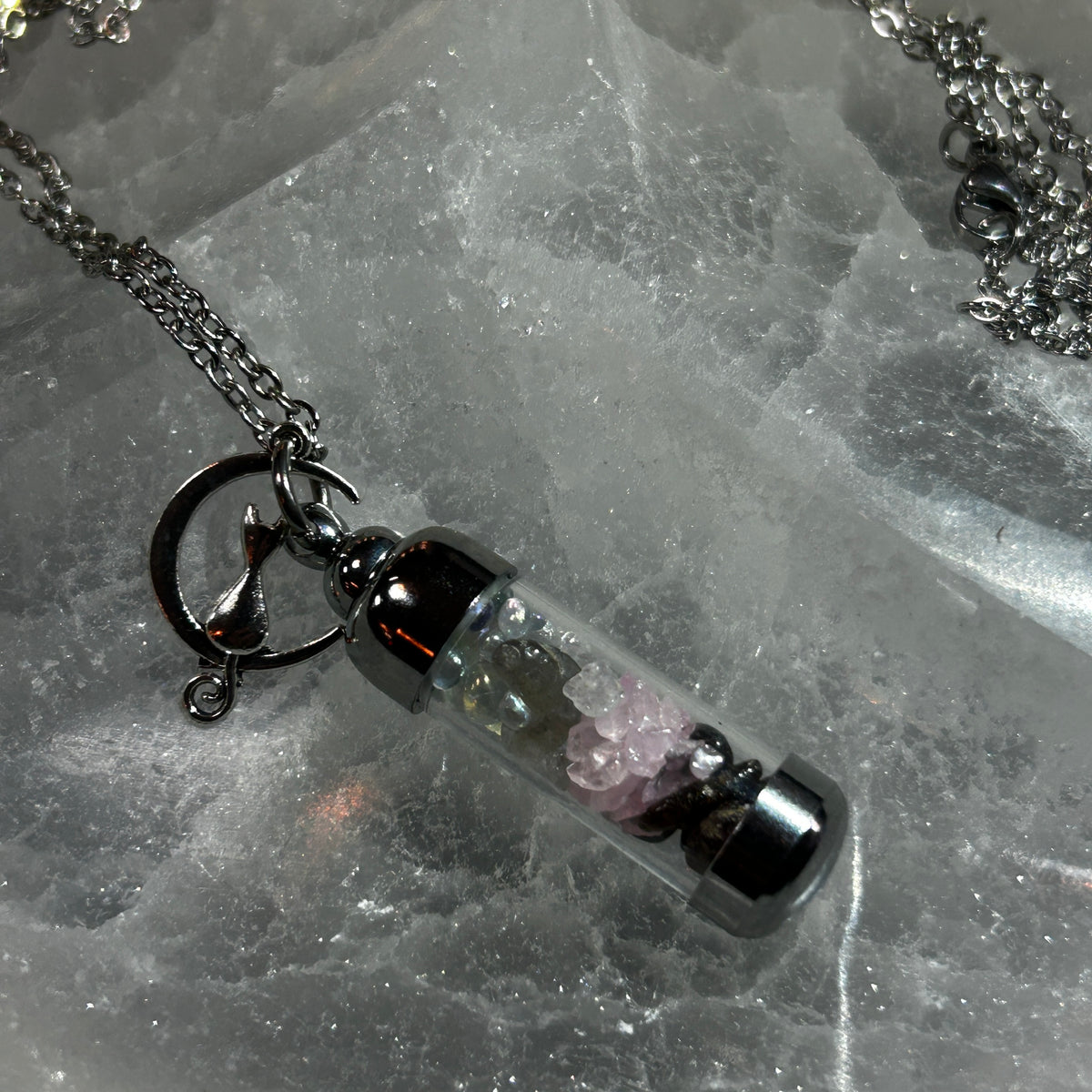 A silver-toned pendant necklace featuring a clear cylindrical vial filled with small pink and dark stones, hanging from a silver chain on a textured gray stone surface.
