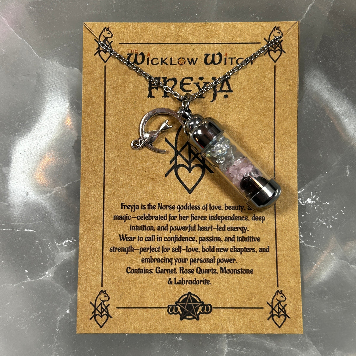 A silver chain necklace with a small clear vial pendant containing pink and black beads, displayed on a brown kraft card labeled 'Wicklow Witch Freya'.