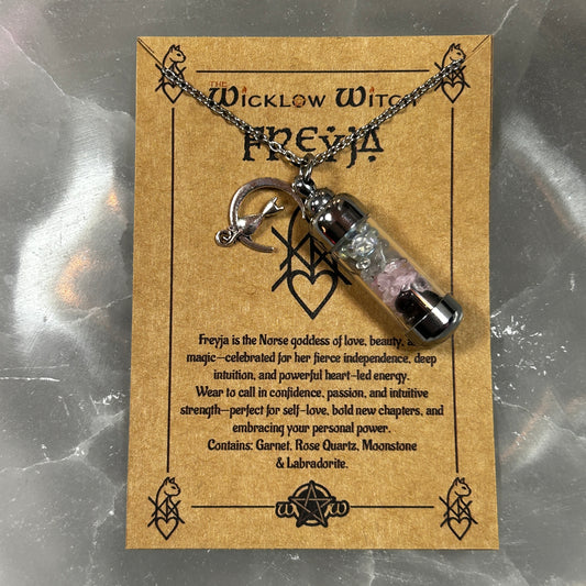 A silver chain necklace with a small clear vial pendant containing pink and black beads, displayed on a brown kraft card labeled 'Wicklow Witch Freya'.