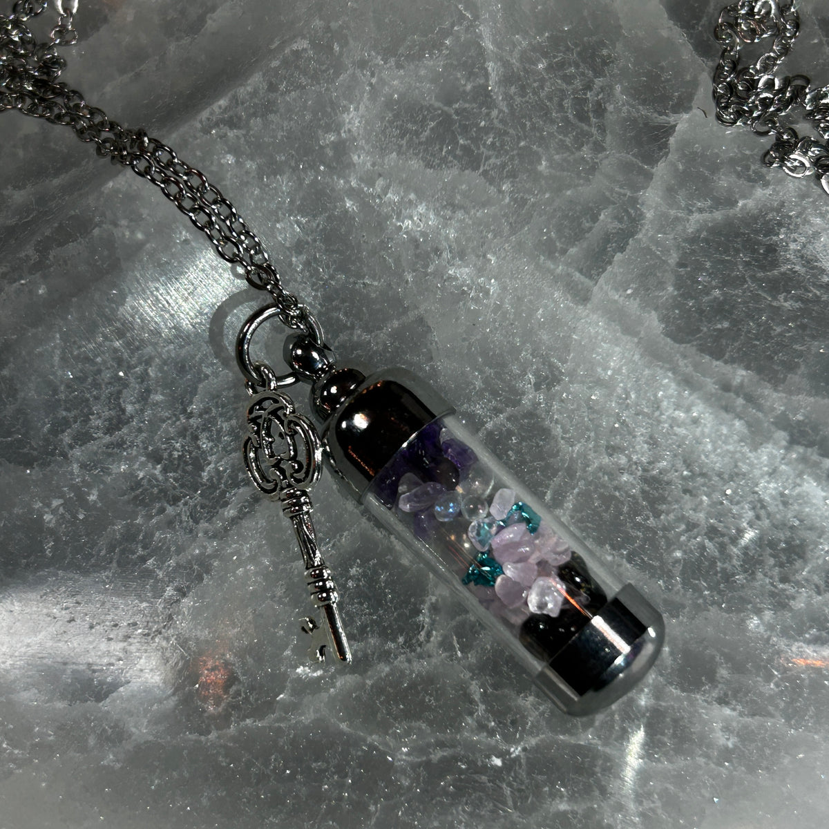 A gothic-style pendant necklace featuring a clear glass vial filled with purple and pink crystals, a silver key charm, and a silver chain, displayed on a textured gray stone surface.