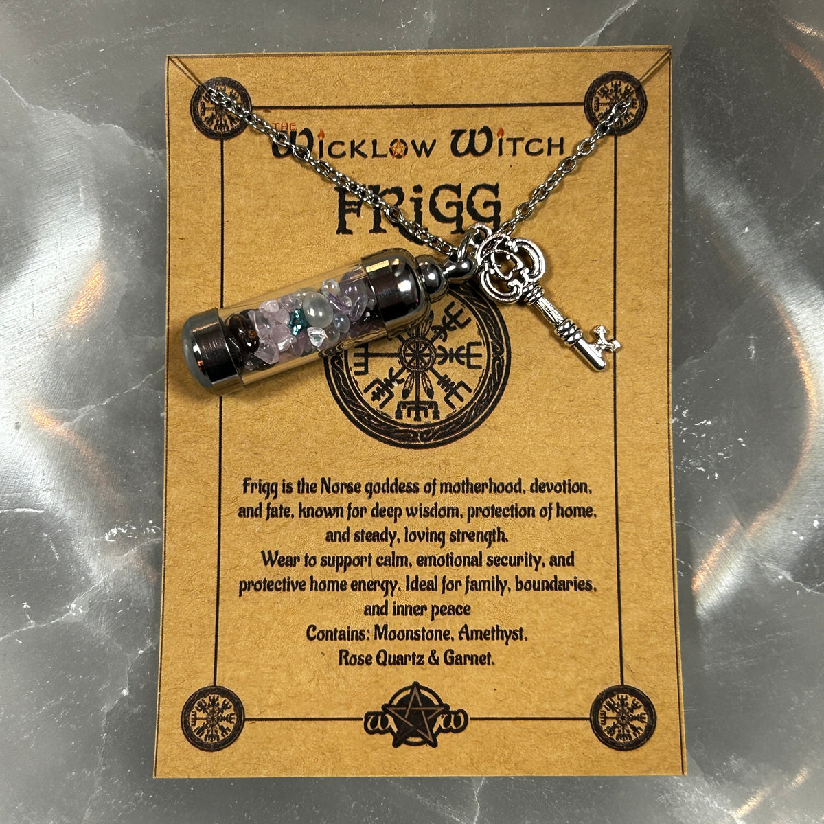 A Frigg necklace with a glass vial containing pink and green crystals, hanging from a silver chain with a key charm, displayed on a brown card labeled 'Wicklow Witch Frigg'.