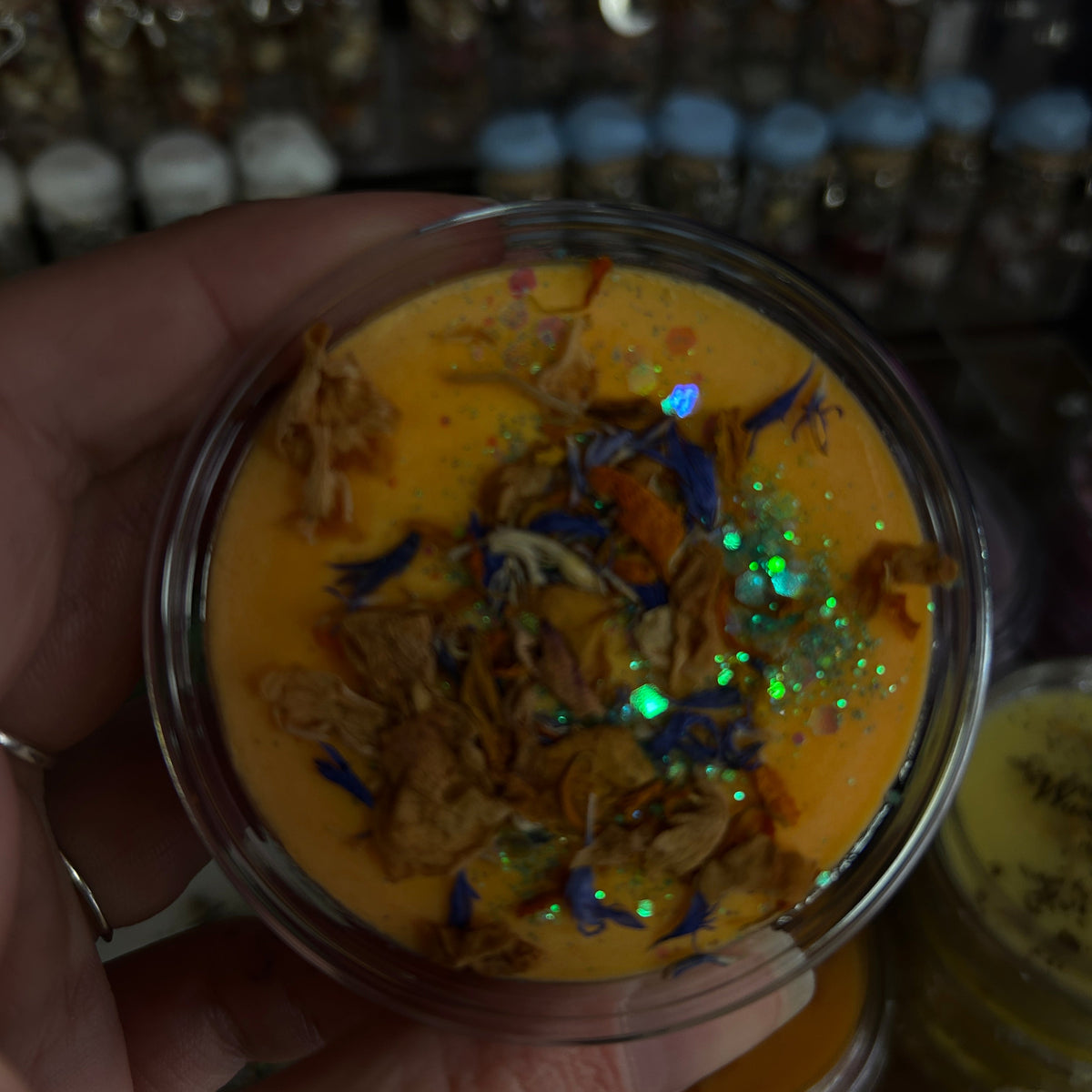 A hand holding a small clear plastic jar filled with bright orange-yellow wax or cream, topped with dried flower petals and iridescent glitter, with blurred bottles in the background.