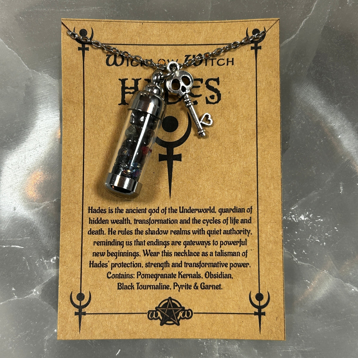 A silver-toned Hades pendant necklace on a black chain, displayed on a brown kraft card with gothic text and symbols. The pendant includes a small vial, key, and skull charm.
