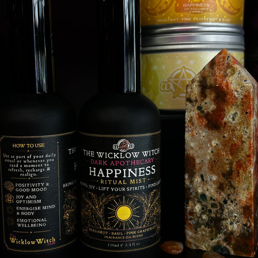 Two bottles of Wicklow Witch Happiness Ritual Mist and a large polished crystal tower, with a stack of metal tins in the background and small polished stones scattered on a black surface.