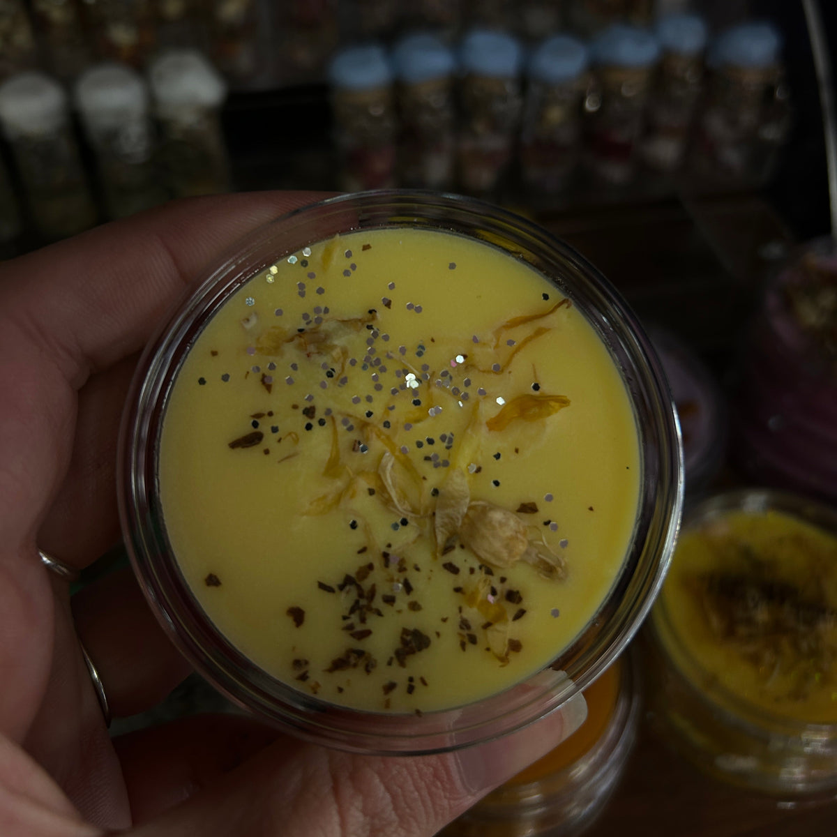 A hand holding a small clear jar filled with a bright yellow wax melt, topped with dried botanical pieces and black seed specks, with other jars blurred in the background.