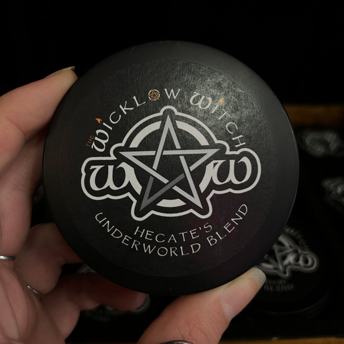 A hand holding a round black cosmetic jar lid with a white pentagram and the text 'The Wicklow Witch' and 'Hecate's Underworld Blend'.