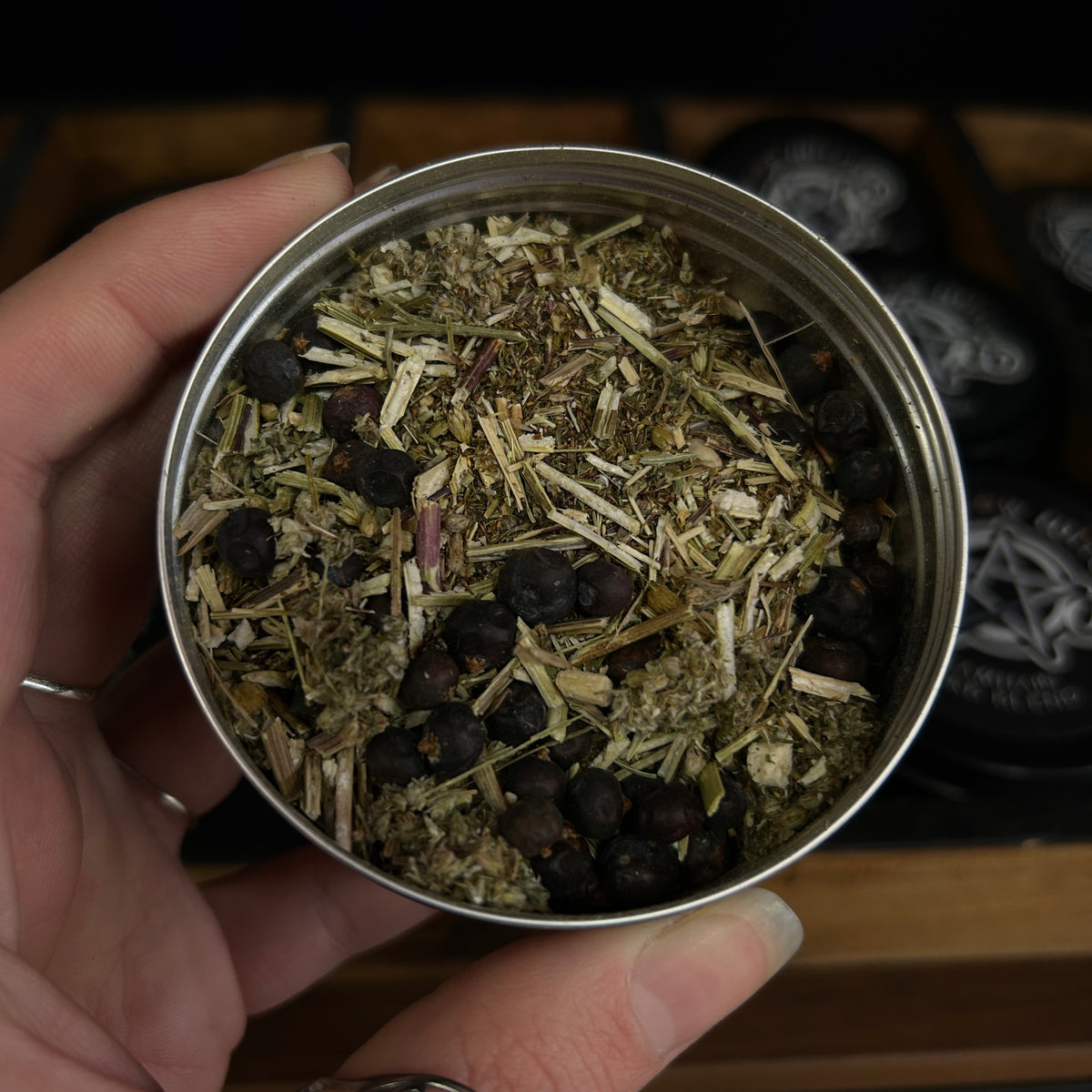 A hand holding the open top of a small silver metal tin filled with dried botanicals and dark berries, with blurred black tins in the background.