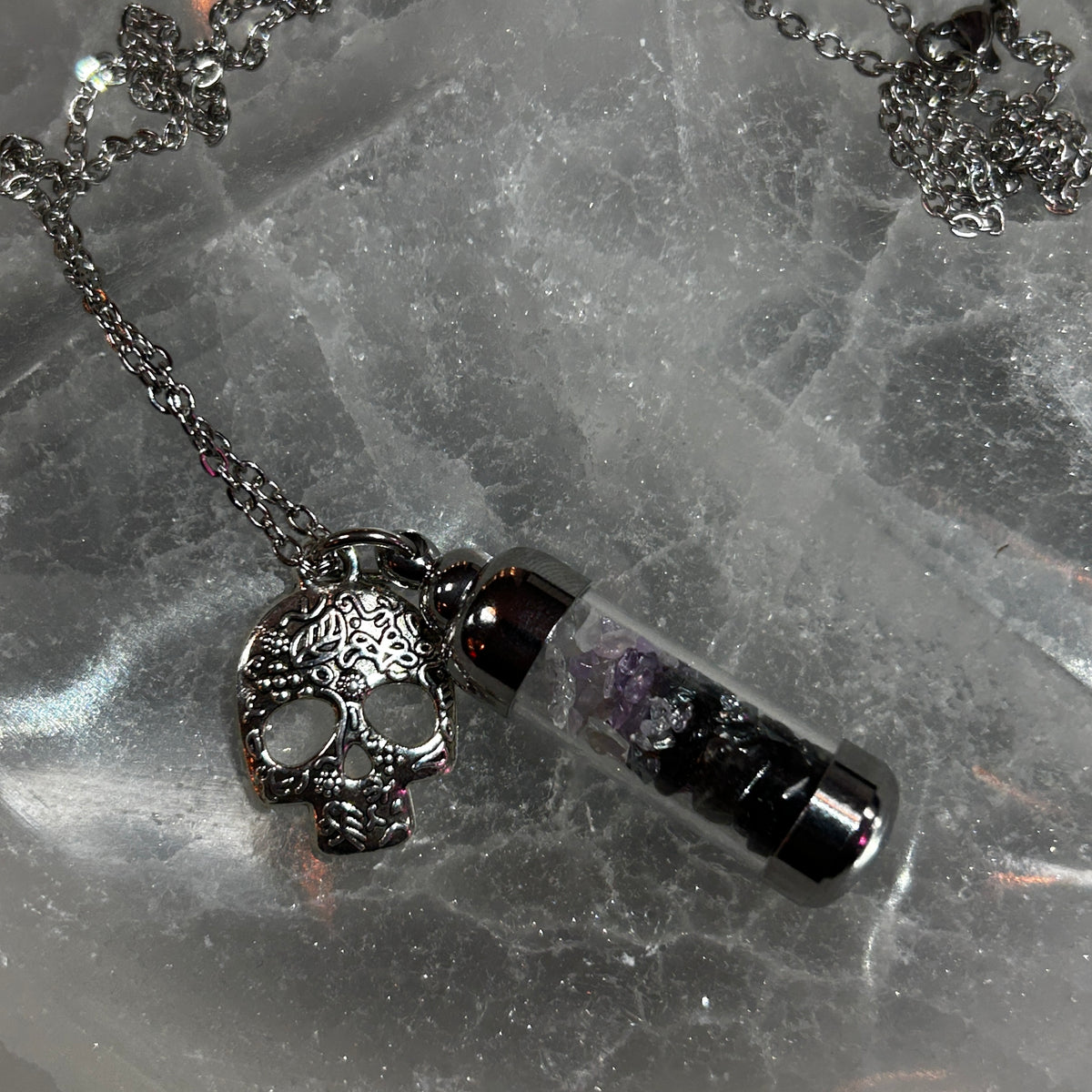A gothic-style silver necklace featuring a skull pendant and a clear cylindrical vial charm filled with dark stones and a purple inlay, displayed on a textured white crystal surface.