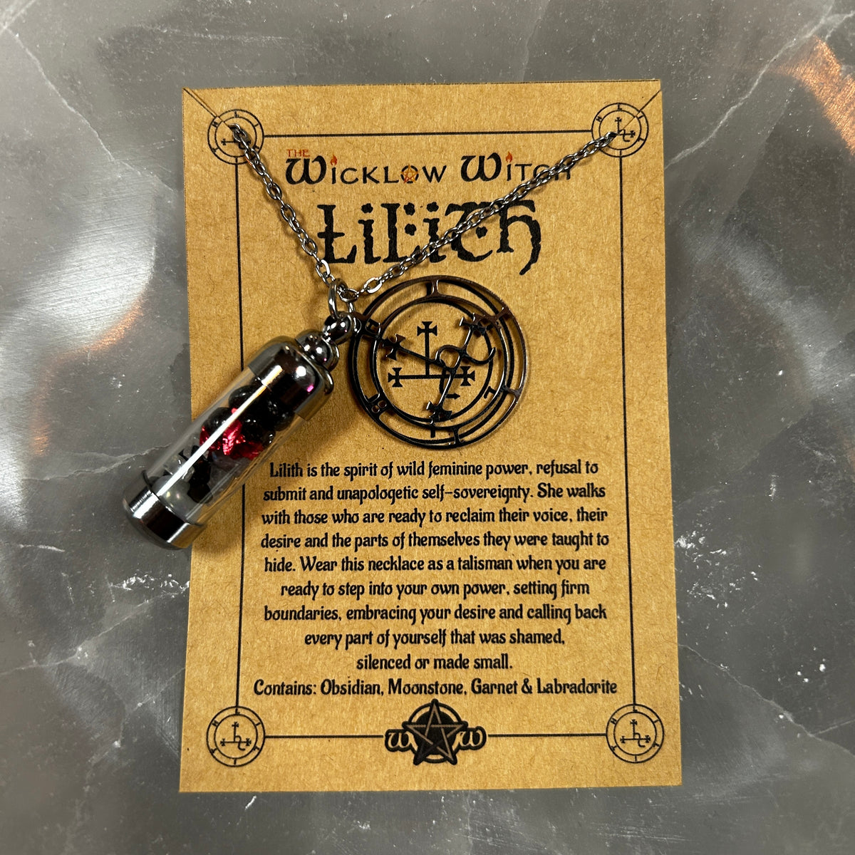 A black glass vial pendant necklace with a red gemstone lies on a brown kraft paper card printed with occult-style text and symbols. The card reads 'Wicklow Witch Lilith' and shows the product as the main subject against a gray marbled surface.
