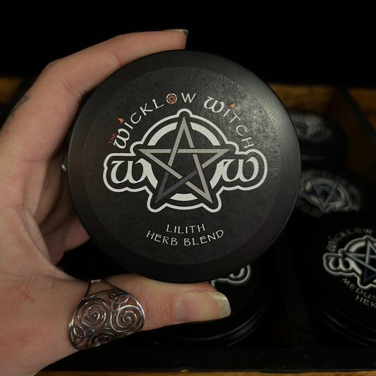 A hand holding a round black tin of Wicklow Witch Lilith Herb Blend, with other matching tins visible in a tray behind it.