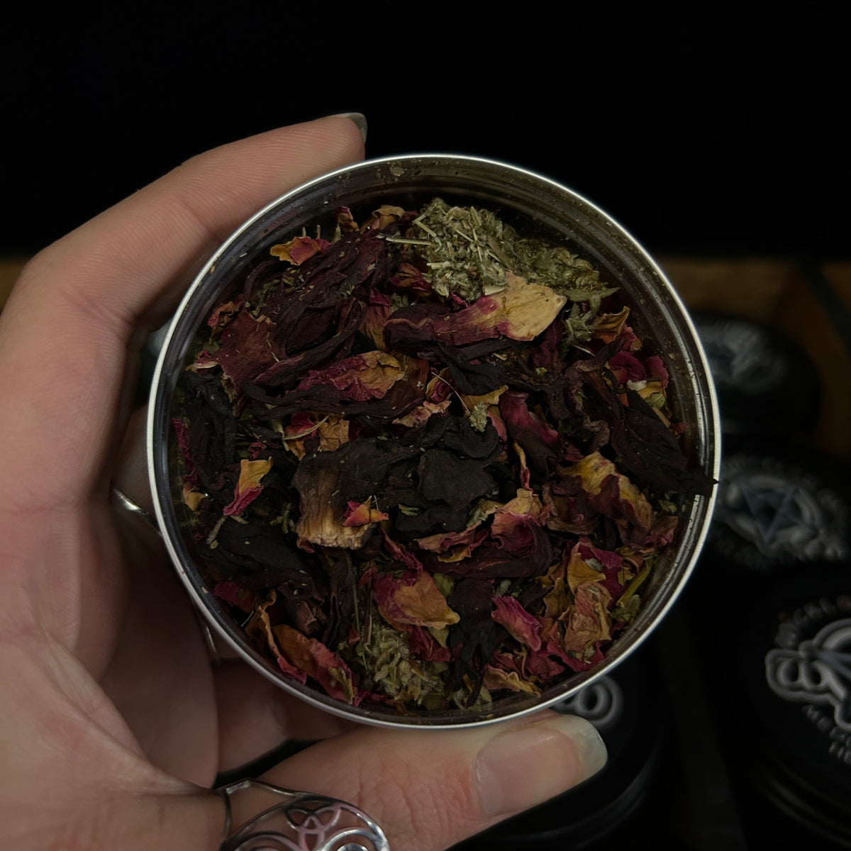 A hand holding a round metal tin filled with dried flower petals and botanical pieces in dark red, purple, brown, and muted green tones, against a black background with other tins partially visible.