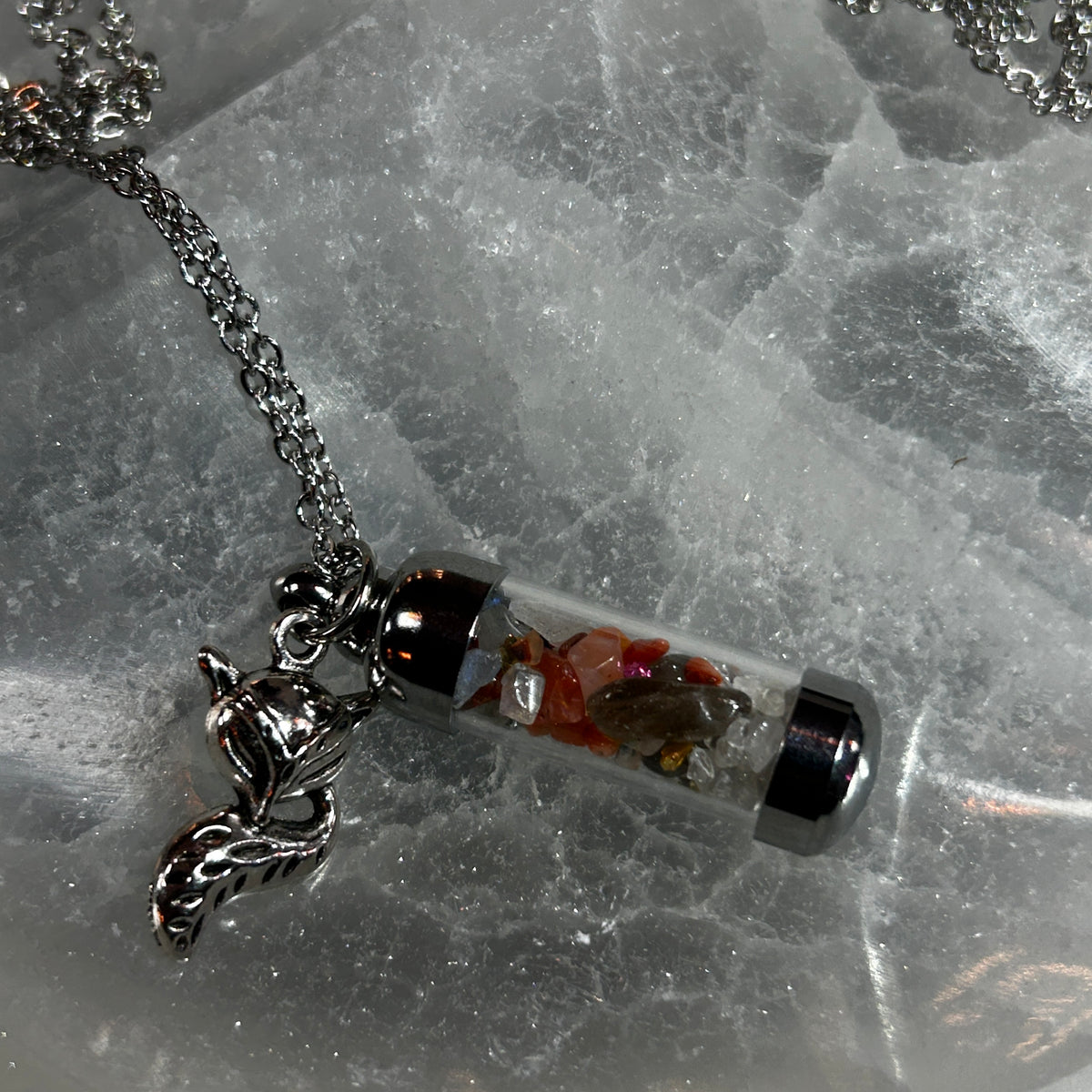 A silver-toned chain necklace with a cylindrical clear glass vial pendant containing small red, orange, and brown stones, displayed on a textured white stone surface.