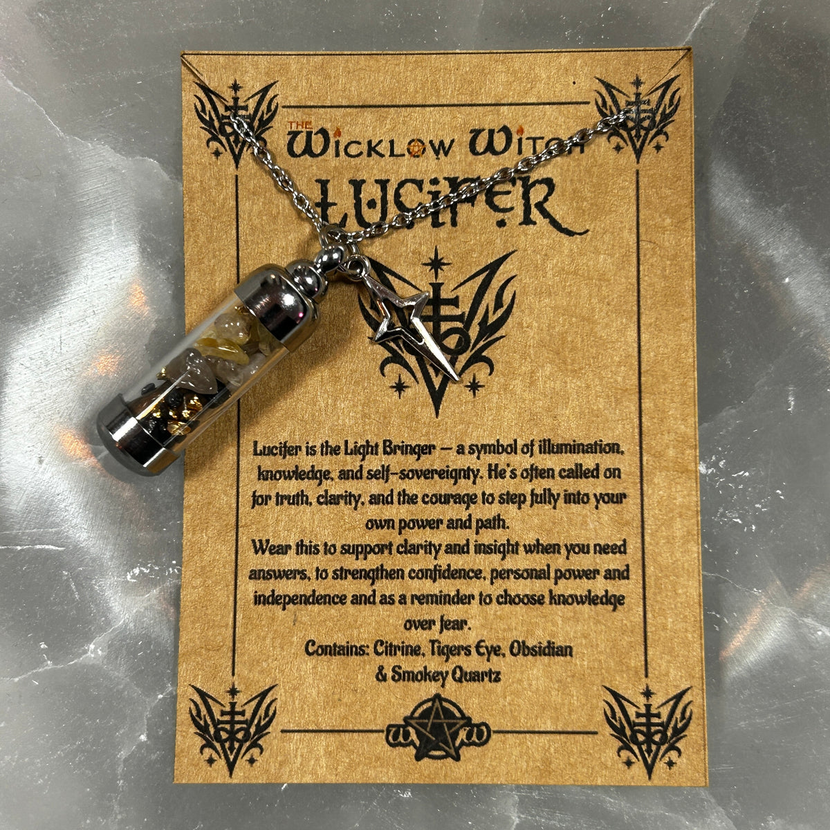 A glass vial pendant necklace with a silver dagger and dried herbs, displayed on its brown backing card titled 'The Wicklow Witch Lucifer'.