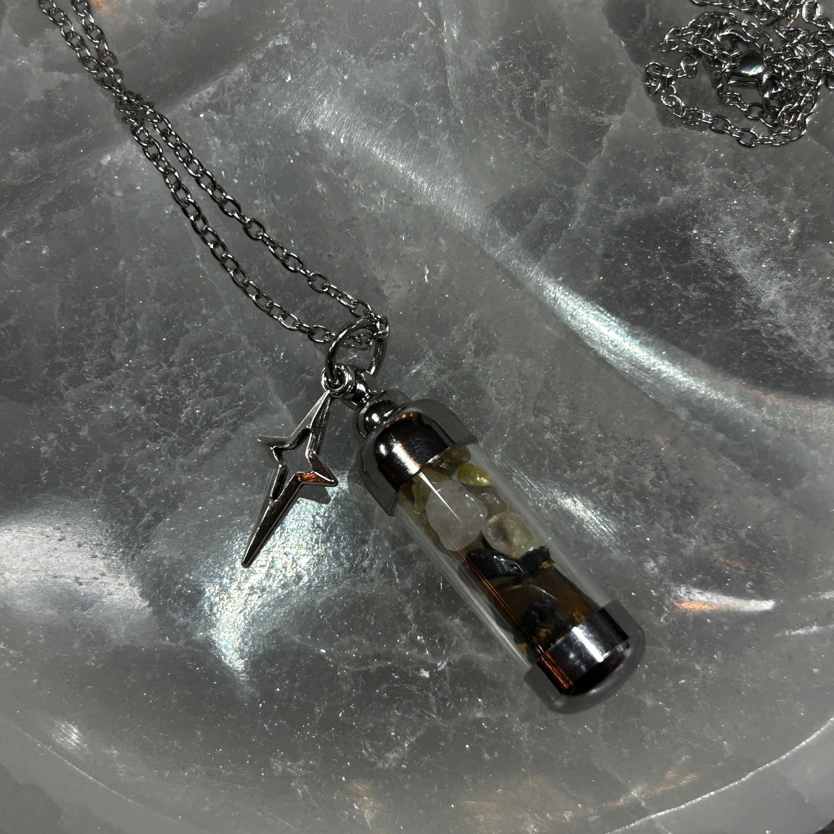 A silver-toned chain necklace with a small clear glass vial pendant containing dried botanical elements and a dangling lightning bolt charm, displayed on a textured gray stone surface.