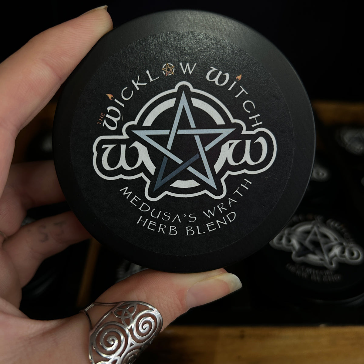 A hand holding a round black tin of 'The Wicklow Witch' 'Medusa's Wrath Herb Blend' against a dark background with other matching tins.