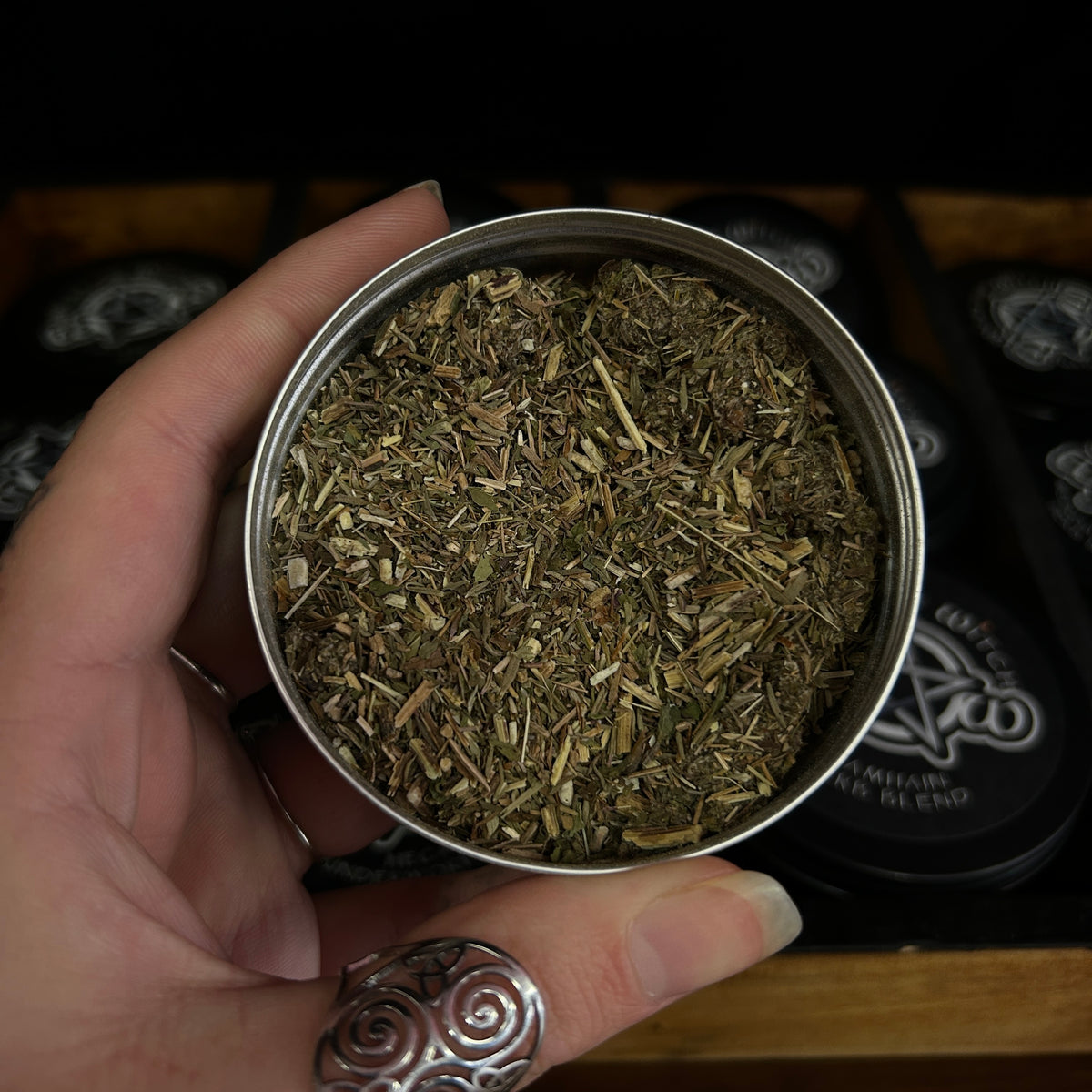 A hand holding a small round metal tin filled with loose dried herbs or tea blend, with black branded tins in the background.