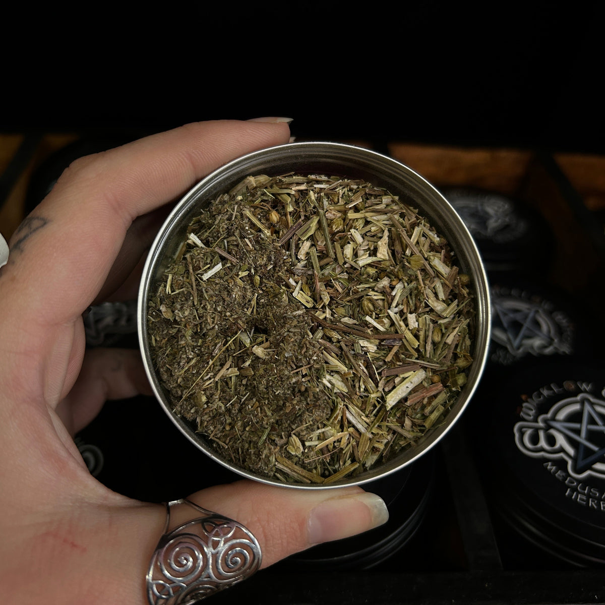A hand holding a small round metal tin filled with dried botanical herbs or tea, with a black tray and branded lids in the background.