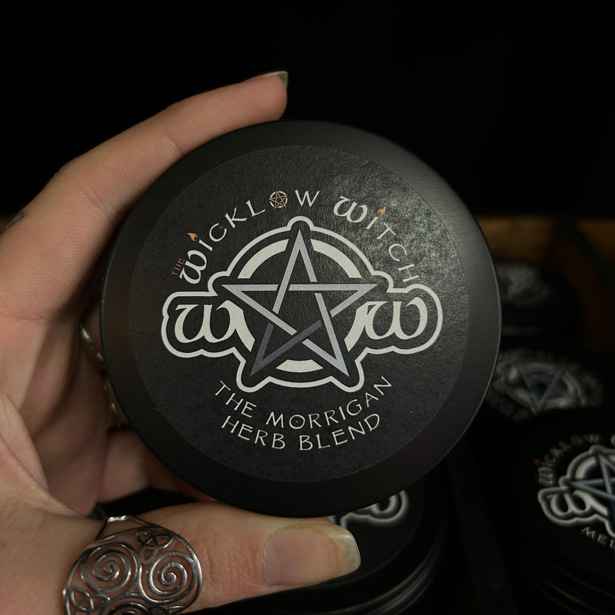 A hand holding a round black tin labeled 'Wicklow Witch The Morrigan Herb Blend' with a pentagram logo, against a dark background with other matching tins visible.