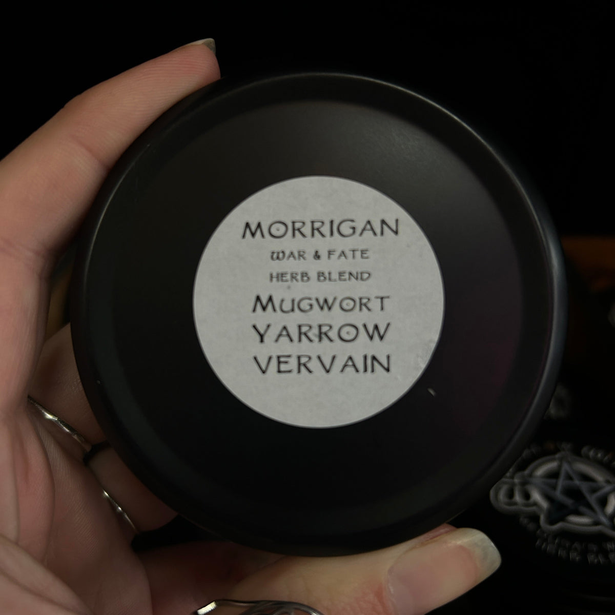 A hand holding a round black tin of Morrigan 'Dark & Fate' herb blend, with a white circular label reading 'Mugwort Yarrow Vervain'.